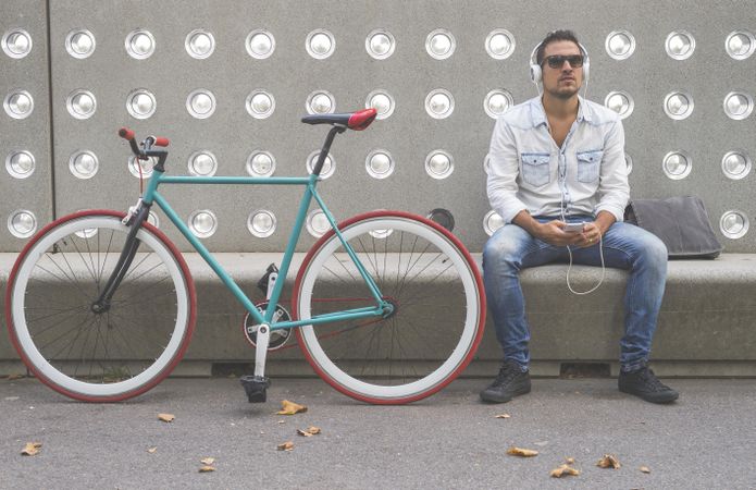 Male listening to music on phone while sitting with bike parked in front of patterned cement wall