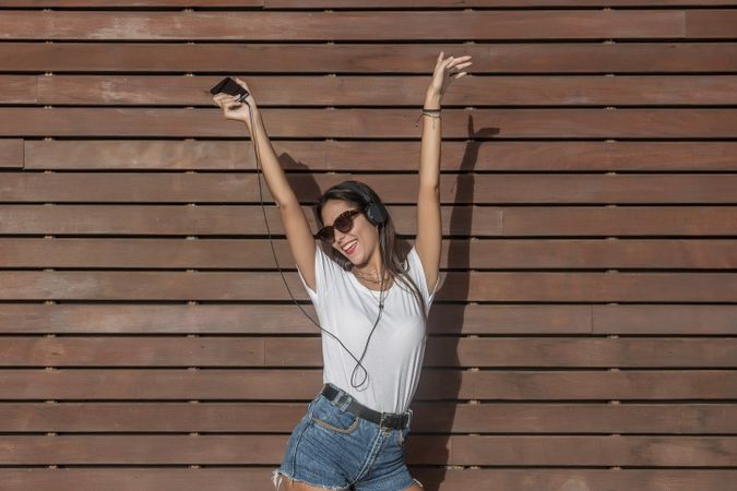 Young woman in light shirt and denim shorts listening to music with headphone and dancing outdoor