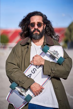 Serious bearded hipster male with skateboard in city park