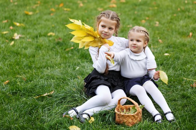 Two young blonde girls sitting in the grass collecting leaves with basket