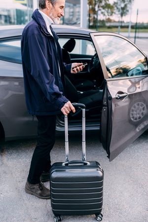 Man about to enter car with suitcase and smartphone in hand, vertical
