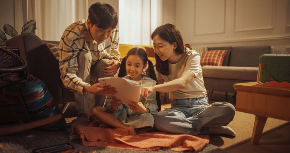 Cute Little Korean Girl Showing Her Drawing To Loving And Happy Mother And Father In Cozy Apartment With Warm Lights.