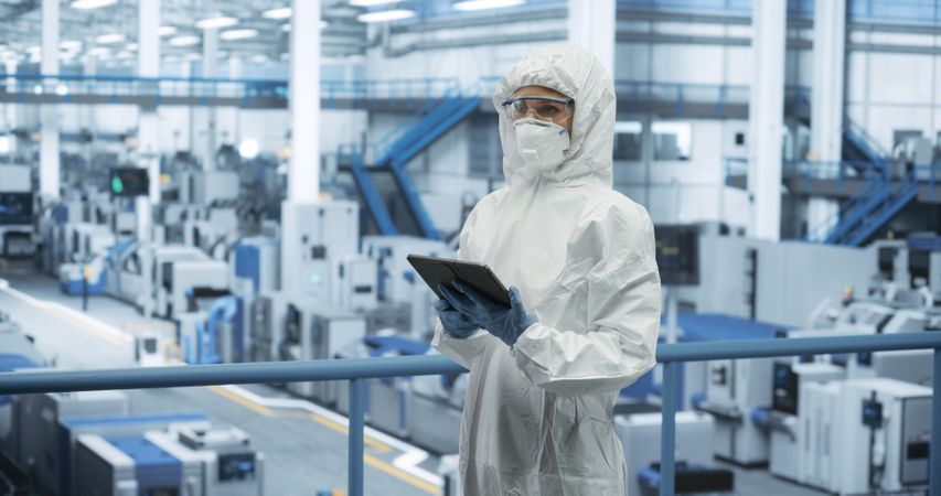 Industrial Scientist Wearing Disposable Protective Suit, Mask and Goggles at a Production Plant. Female Using Tablet Computer, Analyzing Potential Work Hazards and Contamination at a Workplace