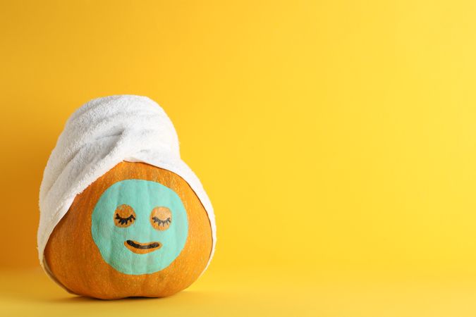 Pumpkin with facial mask and towel on yellow background, copy space