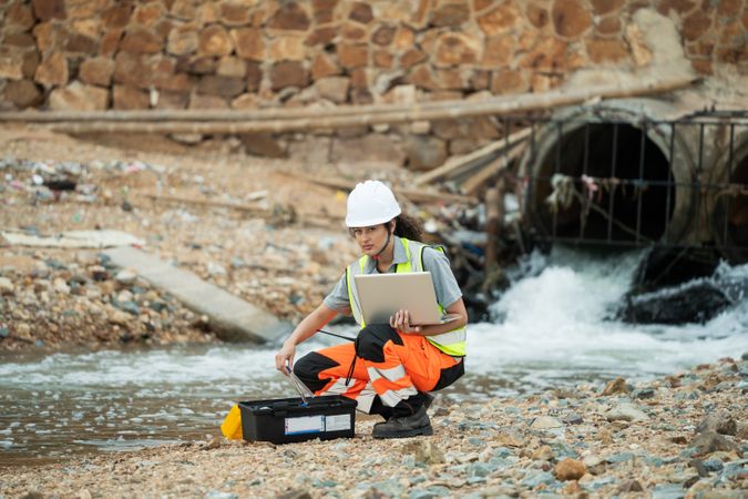 Female engineer with laptop inspecting water drainage pipes at an industrial site