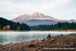Person with fedora hat and backpack sitting on river shore in woods near Mt. Shasta in Siskiyou County, CA, US 0P2la4