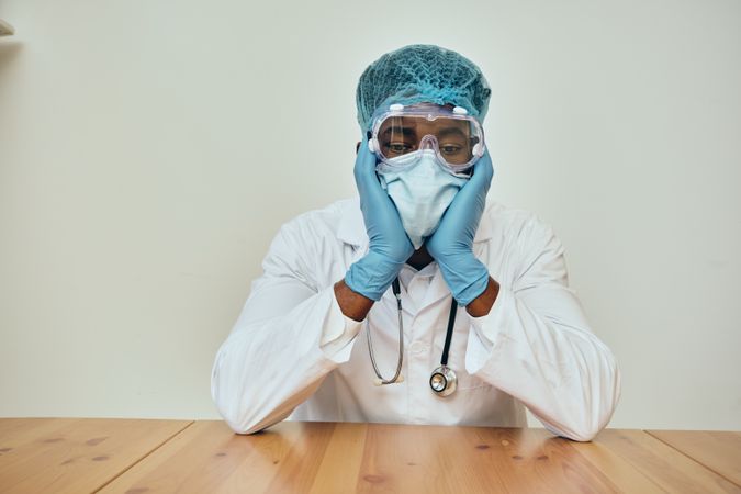 Tired Black male doctor in ppe gear at a table resting his head in his hands