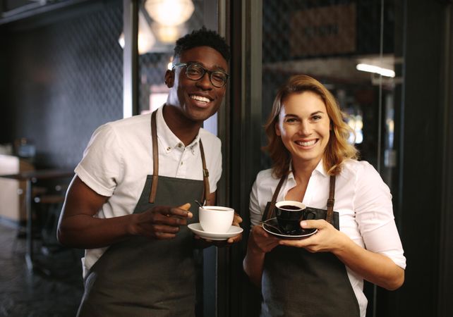 Male and female barista standing at the entrance of coffee shop holding coffee cups