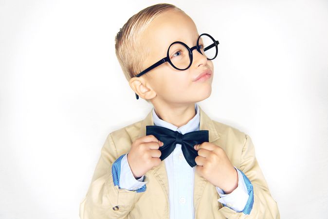 Serious blond boy in glasses fixing his bow tie looking away