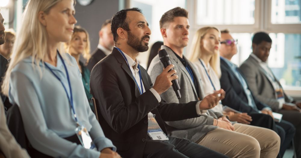 Multiethnic Male Technology Conference Attendee Sitting In Crowd And Using Microphone To Ask A Question Related To Presentation Of Innovative Gadget.