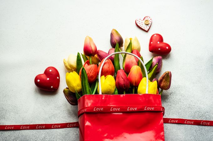 Tulips in shopping bag with gift, red ribbon and heart ornaments on grey counter