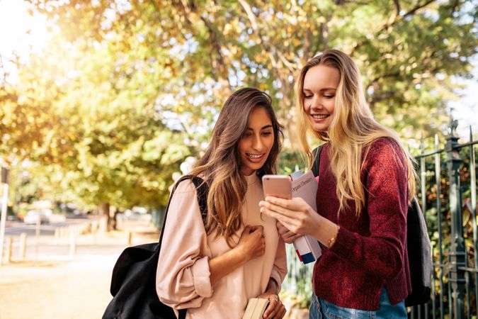 Two female friends smiling while looking at smart phone outdoors