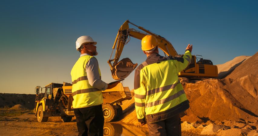 Back View Of White Real Estate Investor With Tablet Computer And Project Manager Talking On Construction Site. Colleagues Wearing Hard Hats And Watching Heavy Machinery Working. Sunny Day
