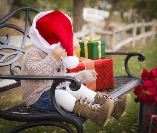 Young Child Wearing Santa Hat Sitting with Christmas Gifts Outside.