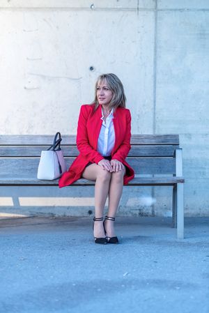 Blonde executive woman wearing red jacket sitting on a city bench while looking away