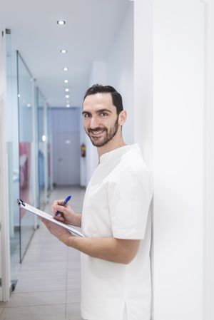 Portrait of a smiling doctor with beard holding a clipboard while leaning on wall in clinic corridor, vertical