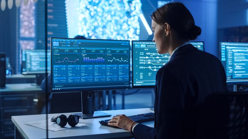 Focused Professional Analyzes Data on Dual Monitors in a Modern Control Room, Optimizing Cybersecurity and System Performance.