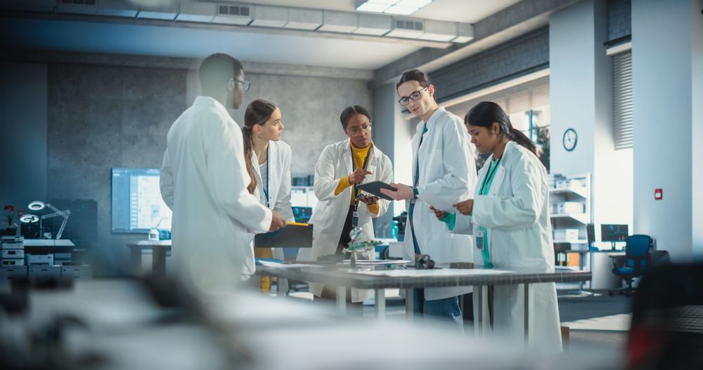 Group of International Multiethnic Lab Specialists Gathering Around a Table to Discuss Blueprints and Components for a High Technology Robotic Project