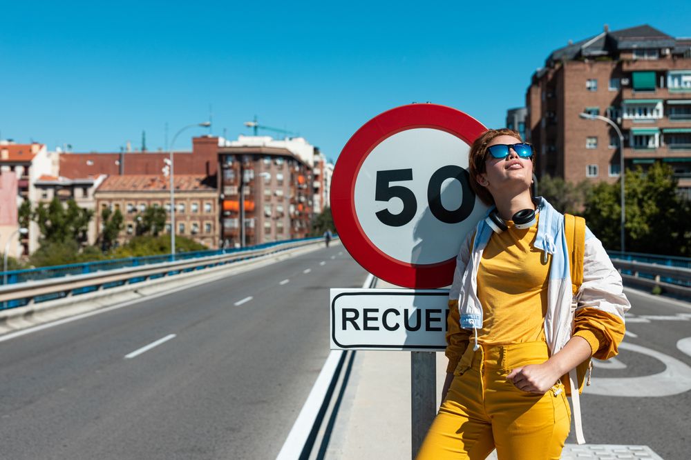 Trendy joyful young woman in sunglasses enjoying view beside road sign on bridge in sunny day