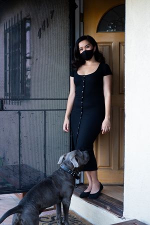 Woman standing by front door in dark dress and mask looking at camera