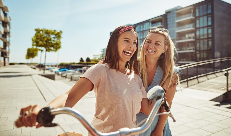 Two laughing female friends with arms on each others shoulders while walking with bike, close up