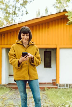 Smiling woman using smartphone near countryside house