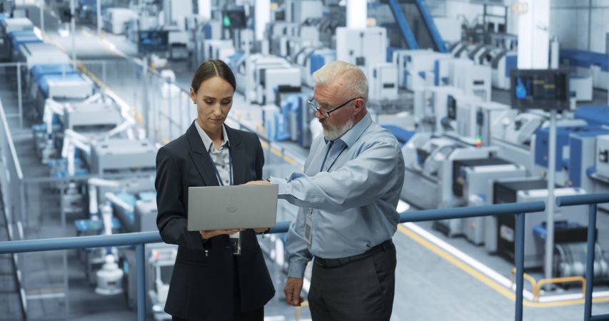 Mature Engineer and a Female Technician Having a Conversation and Using Laptop Computer. Specialists Monitoring, Analyzing Conditions at a Modern Electronics Factory with Automated Robotic Arms