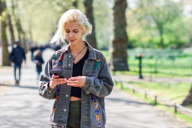 Blonde woman in jean jacket looking down at messages on cell phone while walking in park