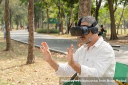 Mature grey haired man sitting outside in park with hands up wearing VR headset 4Mm8r4
