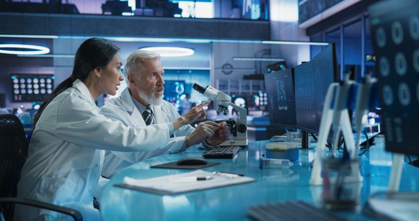 Biotechnology Medical Research Center: Female Asian Neuroscientist Using Desktop Computer And Talking To Male Doctor With Microscope.