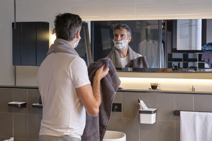 Confident male with shaving foam on face looking at mirror