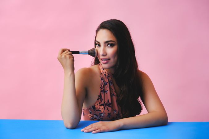 Hispanic woman with long brown hair holding large make up brush and looking at camera