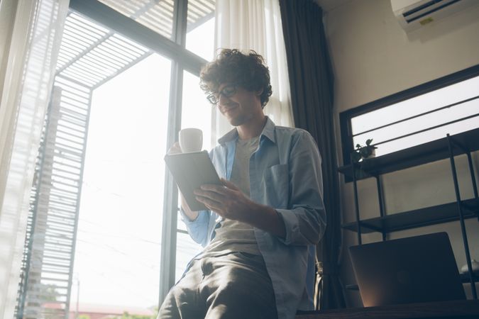 Man working from home perched on desk reading tablet sipping coffee