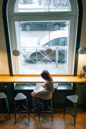 Side view of woman sitting on stool chair reading a book near arched window