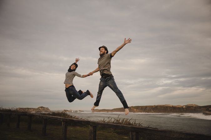Couple holding hands and jumping on bench on shore near water