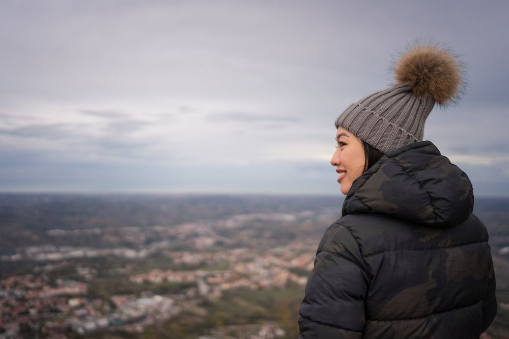 Relaxed Asian woman in warm hat with pompom sightseeing and looking at city down