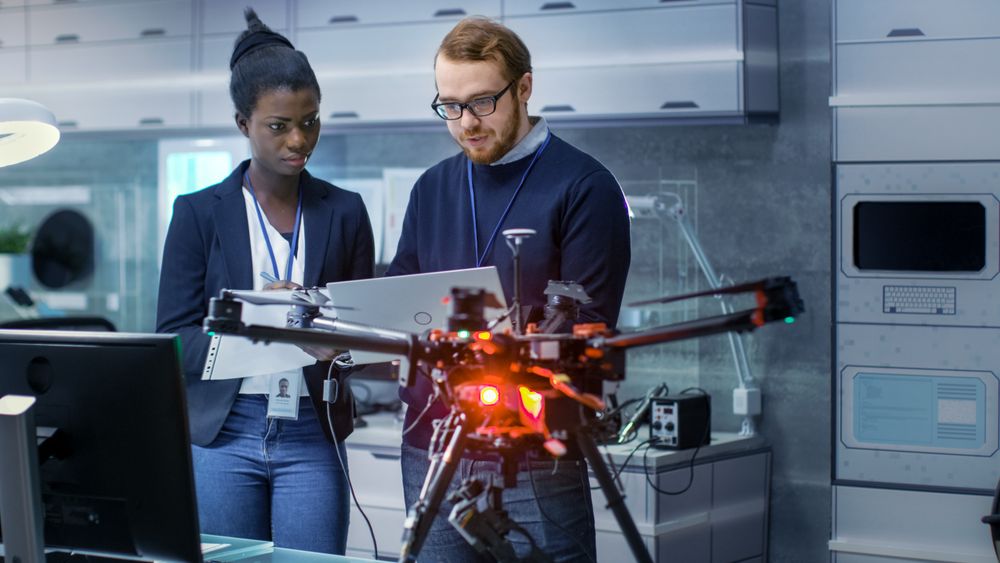 White Male and Black Female Engineers Working on a Drone Project with Help of Laptop and Taking Notes.
