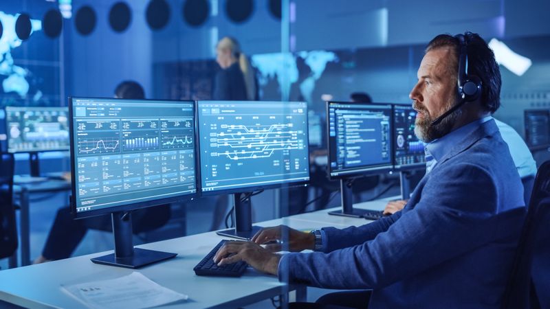 Bearded Analyst in Blue Shirt Monitors Data on Multiple Screens in Dimly Lit Command Center.
