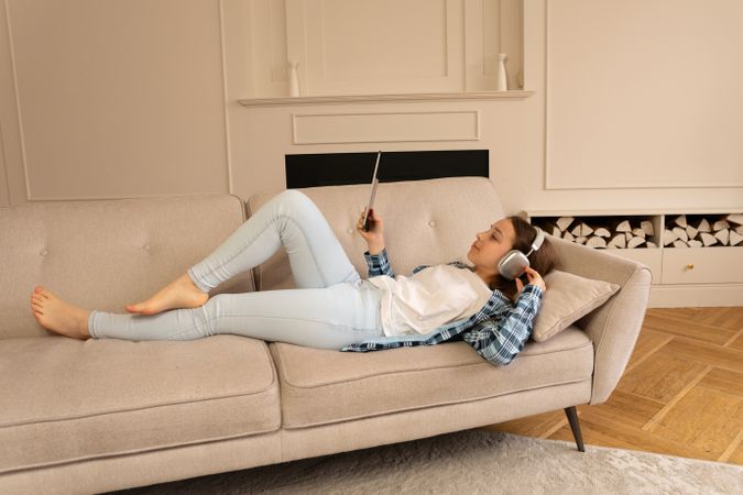 Girl lying on sofa wearing headphones while using tablet.