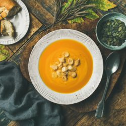 Pumpkin soup with croutons, bread, seeds, spoon on wooden table with leaves, square crop