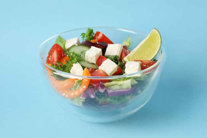 Glass bowl with fresh salad on blue background, space for text