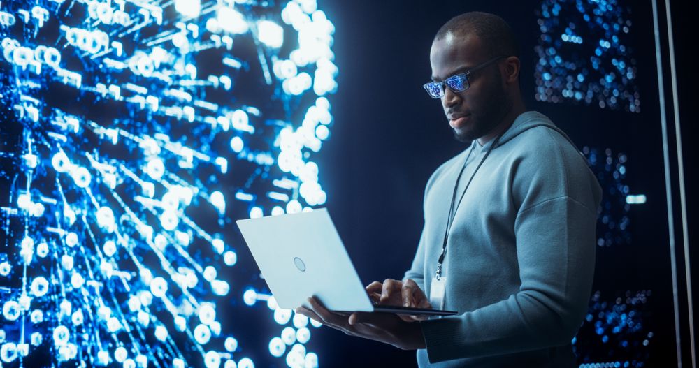 Portrait of Young Black Man Working on Laptop Computer, Looking at Big Digital Screen Displaying Neural Network Visualisation