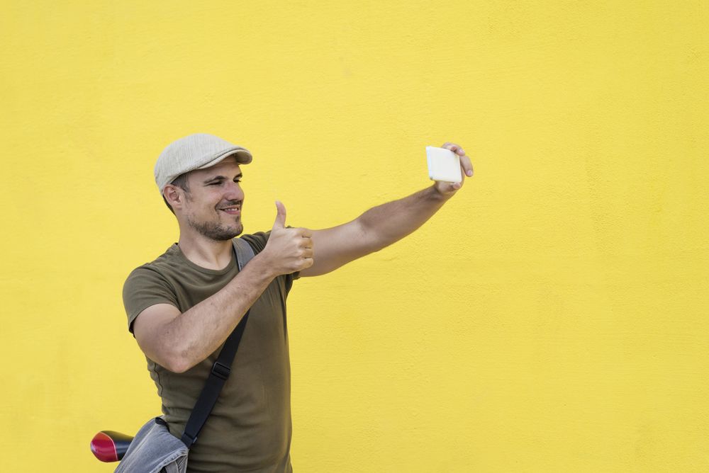 Male in hat and sunglasses taking selfie giving thumbs up on phone while sitting on bicycle