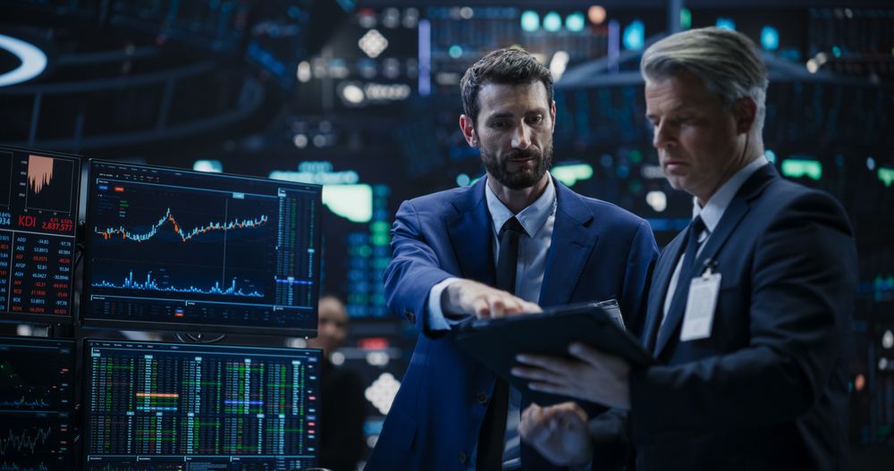 Team of Two Handsome Stock Exchange Brokers Using Tablet Computer, Chatting About the Asset Management Strategy for an International Business Client, Working in a Office Building with Computer Screens