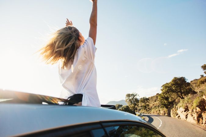Rearview of woman standing out of sunroof on sunny day and celebrating