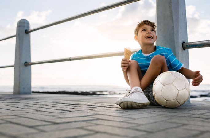 Happy boy eating an ice cream sitting on a pier with soccer ball