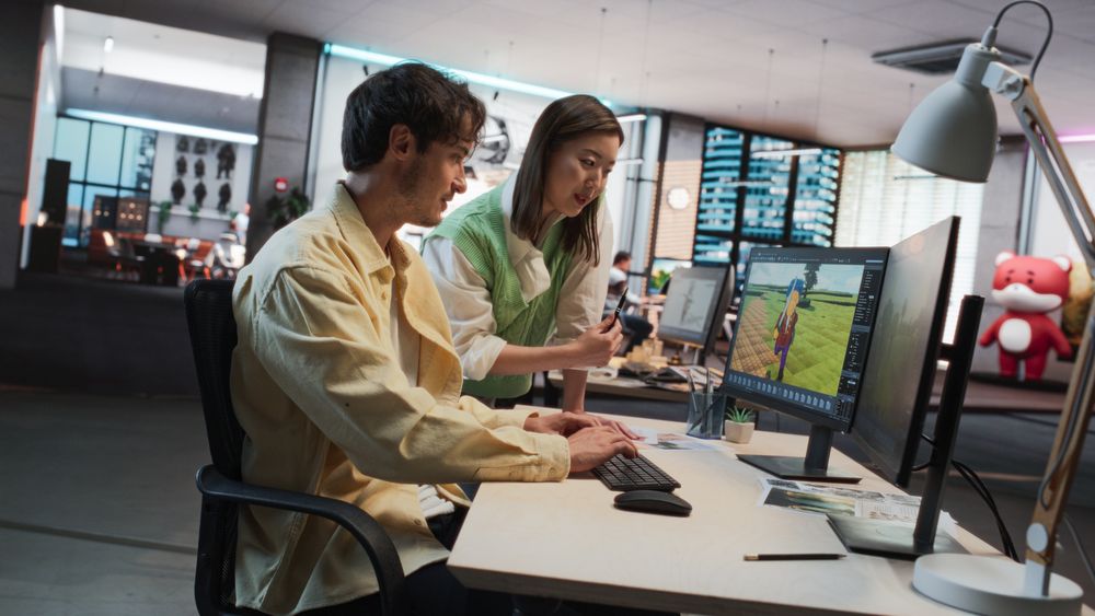 Male Game Designer Using Desktop Computer With 3D modelling Software And Talking to Asian Female Art Director in Game Development Studio Office.