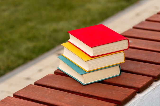 Three colorful books stacked horizontally on park wooden bench.