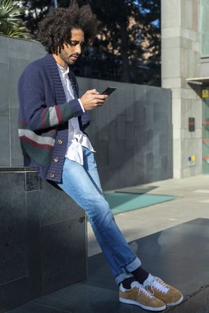 Young Black man using his smartphone while leaning on wall on sunny day