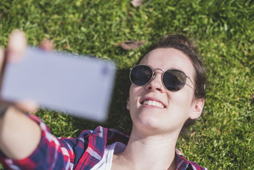 Above view of a young smiling hipster woman lying on grass in a sunny day at a park while taking a selfie with a mobile phone